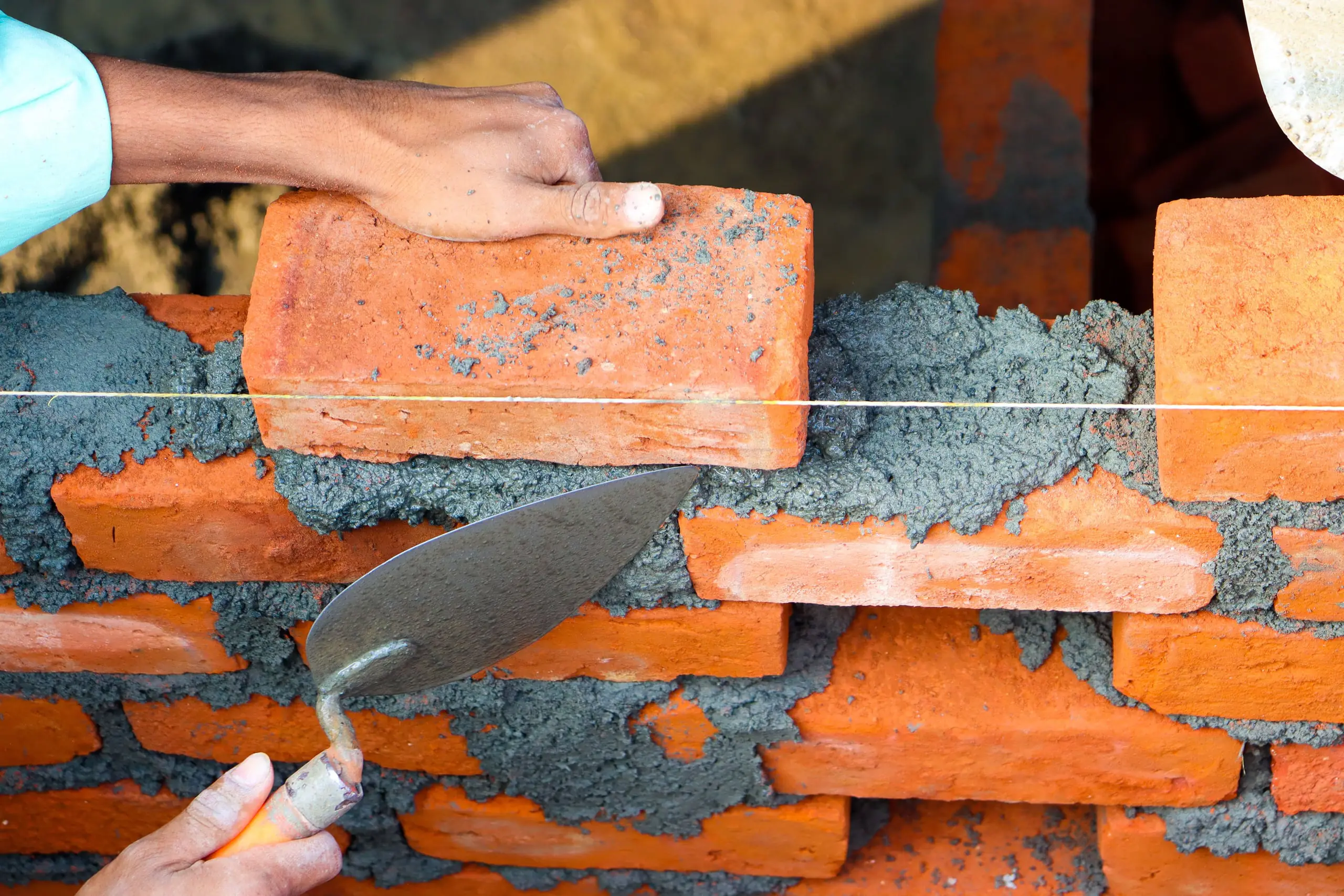 A bricklayer setting a red brick on fresh mortar along a string line, trowel in hand