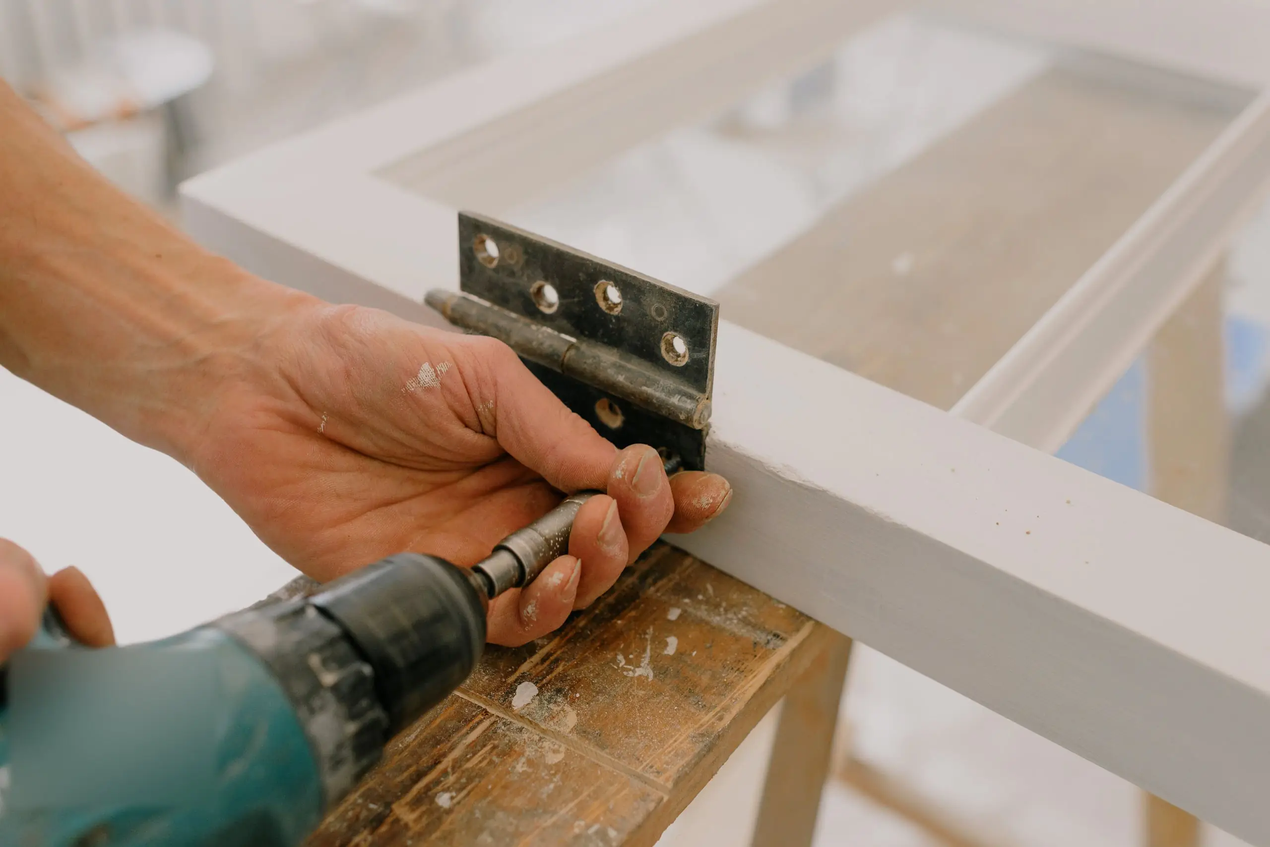 Hands fixing a metal hinge to a white window frame with a cordless drill