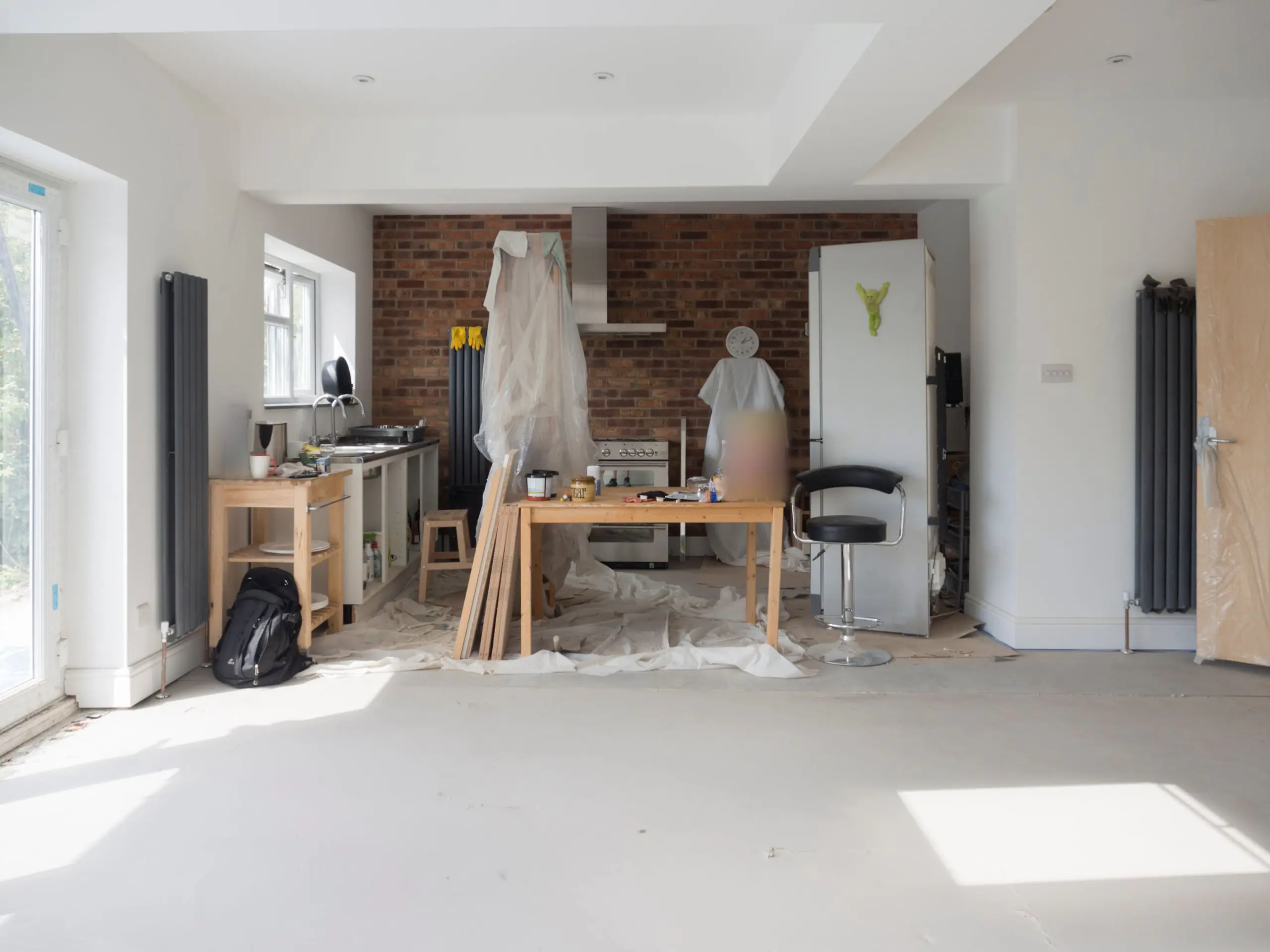 A kitchen and living space mid-refurbishment, with dust sheets, a bare floor and a brick feature wall